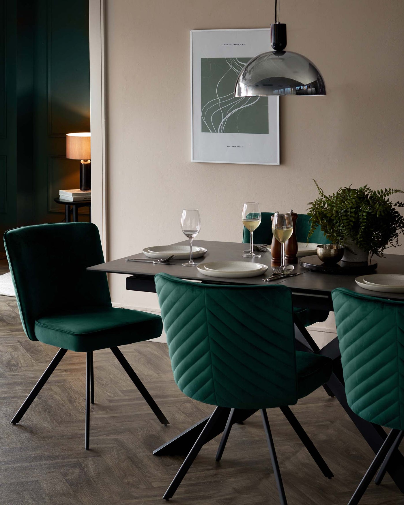 Modern dining room featuring a sleek black rectangular table with matching angular black legs, complemented by four luxurious emerald green upholstered chairs with a distinctive chevron quilting pattern on the backrest. The chairs showcase a plush seat and angled metal legs that mirror the table's design. A contemporary silver dome pendant light hovers above the scene, adding a metallic accent to the sophisticated colour palette.