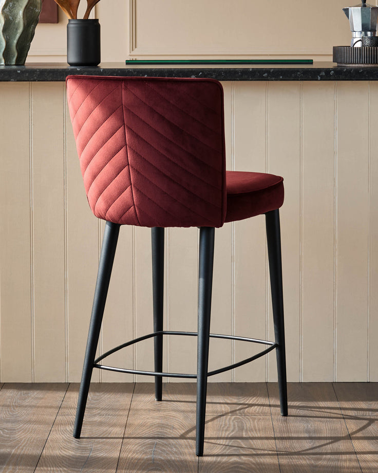 Burgundy velvet bar stool with black legs and silver footrest, positioned in front of a beige panelled wall.