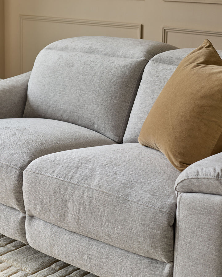 Serene 2-seater fabric recliner sofa in light grey with curved arms, plush cushions, and a tan decorative pillow.