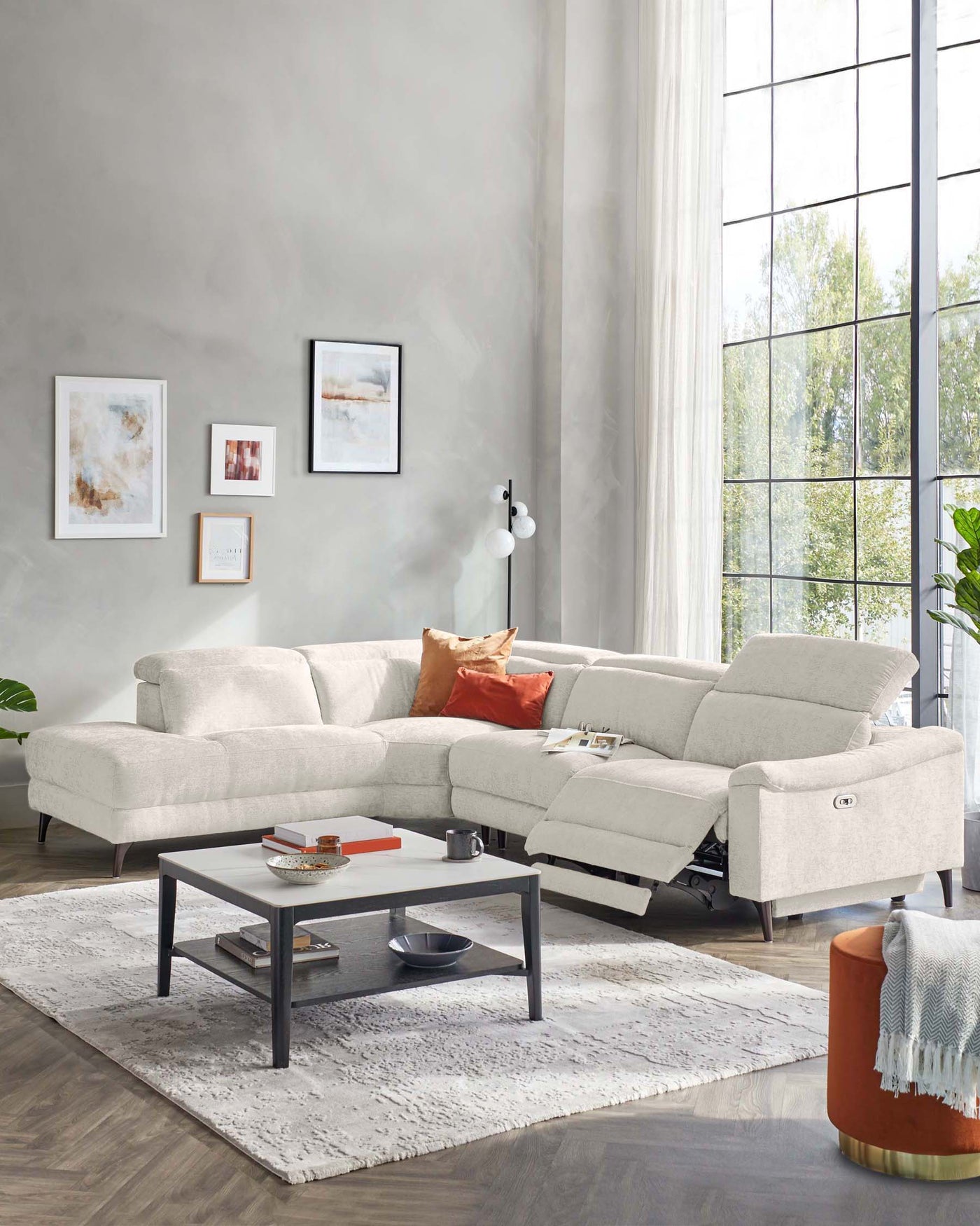 Modern light grey sectional sofa with chaise and reclining function, situated on a white plush area rug. A rectangular black metal coffee table with a grey surface, featuring a lower shelf, is centred in front of the sofa. An orange cylindrical ottoman with a gold base is positioned to the side, adorned with an off-white fringe throw blanket.