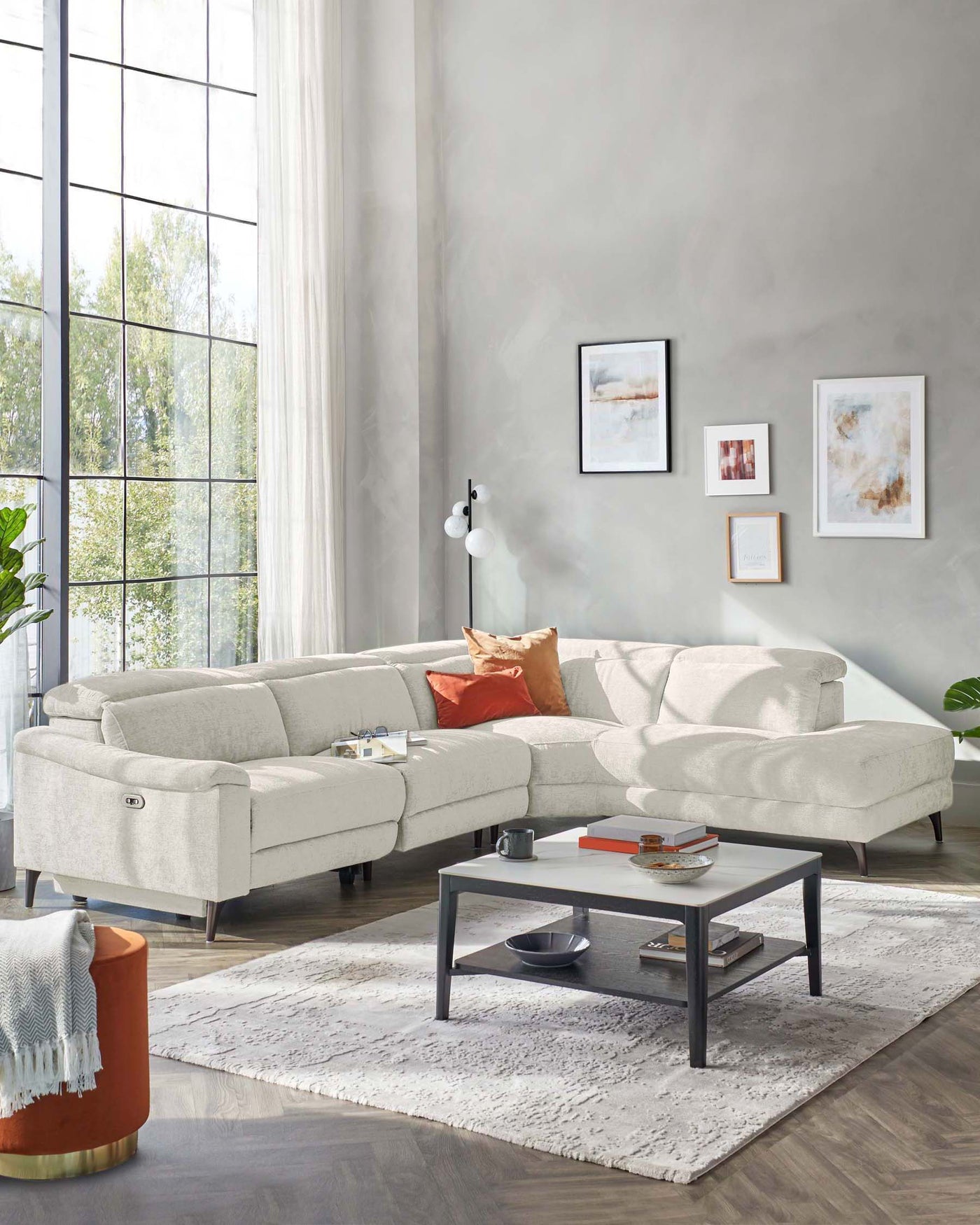 Modern light grey sectional sofa with chaise on the left and plush back cushions, paired with a geometric black metal and wood coffee table on a soft textured area rug. In the corner, a round burnt orange side table with a metallic base complements the setting.