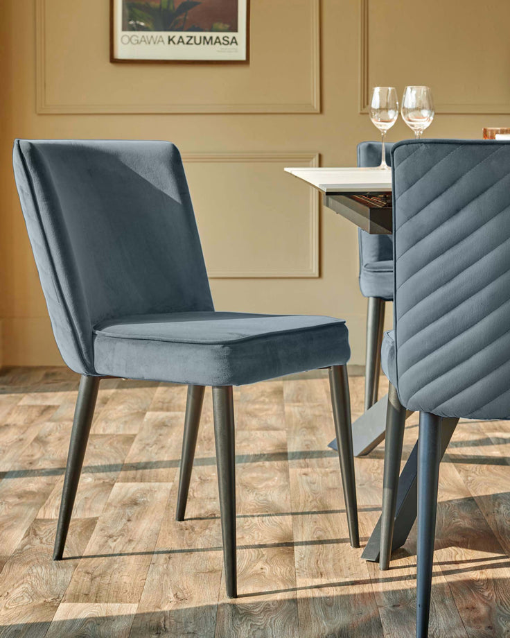 Two modern dining chairs in blue velvet with black legs, positioned around a dining table on a wooden floor.