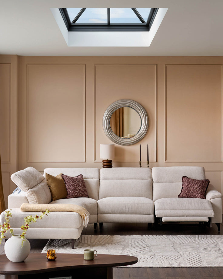 Kyla round mirror with silver finish displayed on a beige wall above a cream sectional sofa in a modern living room.