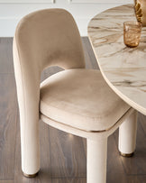 Shay dining chair in champagne velvet with cylindrical legs and brass foot caps, shown at marble table.