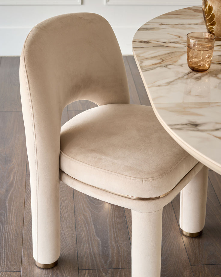 Shay dining chair in champagne velvet with cylindrical legs and brass foot caps, shown at marble table.