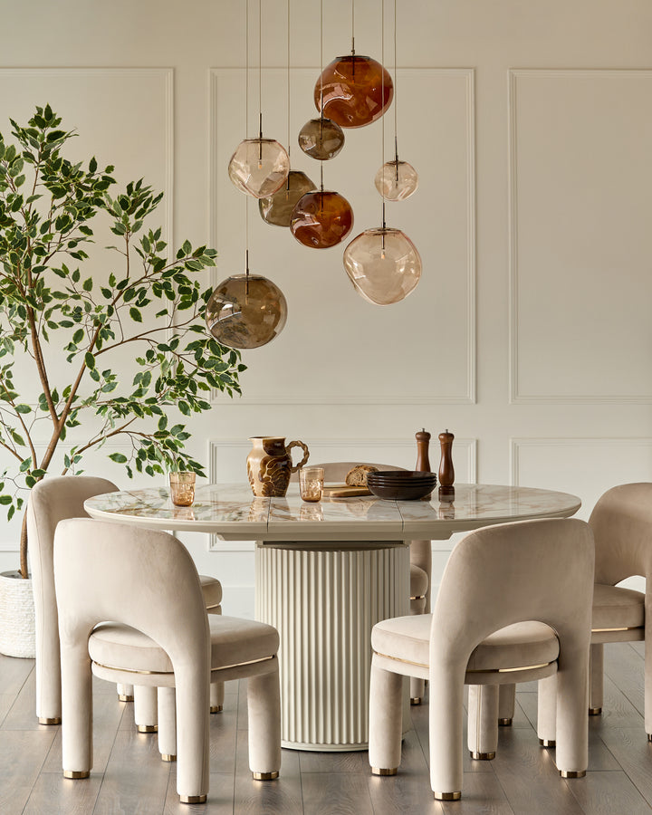 Shay dining chair in champagne velvet with curved back and cylindrical legs in a cream dining room setting.