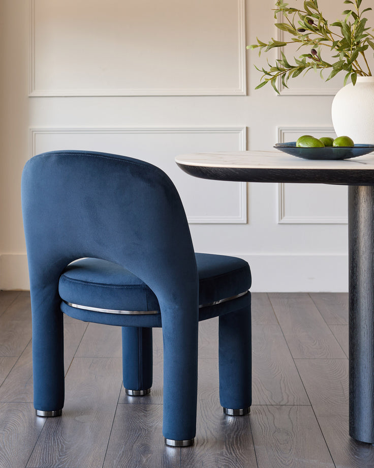 Dark navy velvet dining chair with curved back and cylindrical legs with metal caps, positioned at a round table.