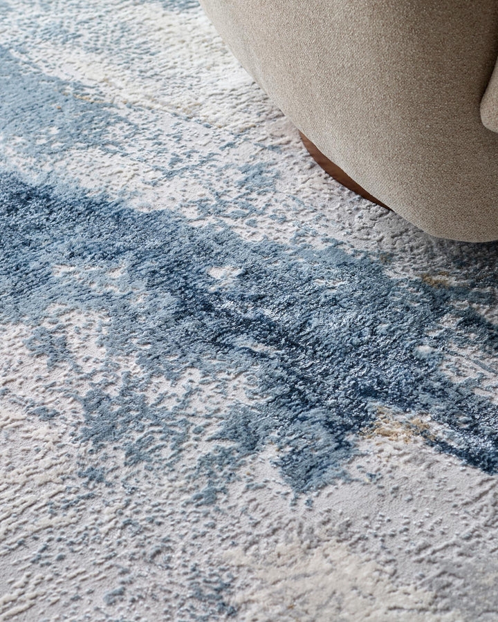Neutral fabric armchair shown partially on a textured, blue and white rug, creating a cosy interior scene.