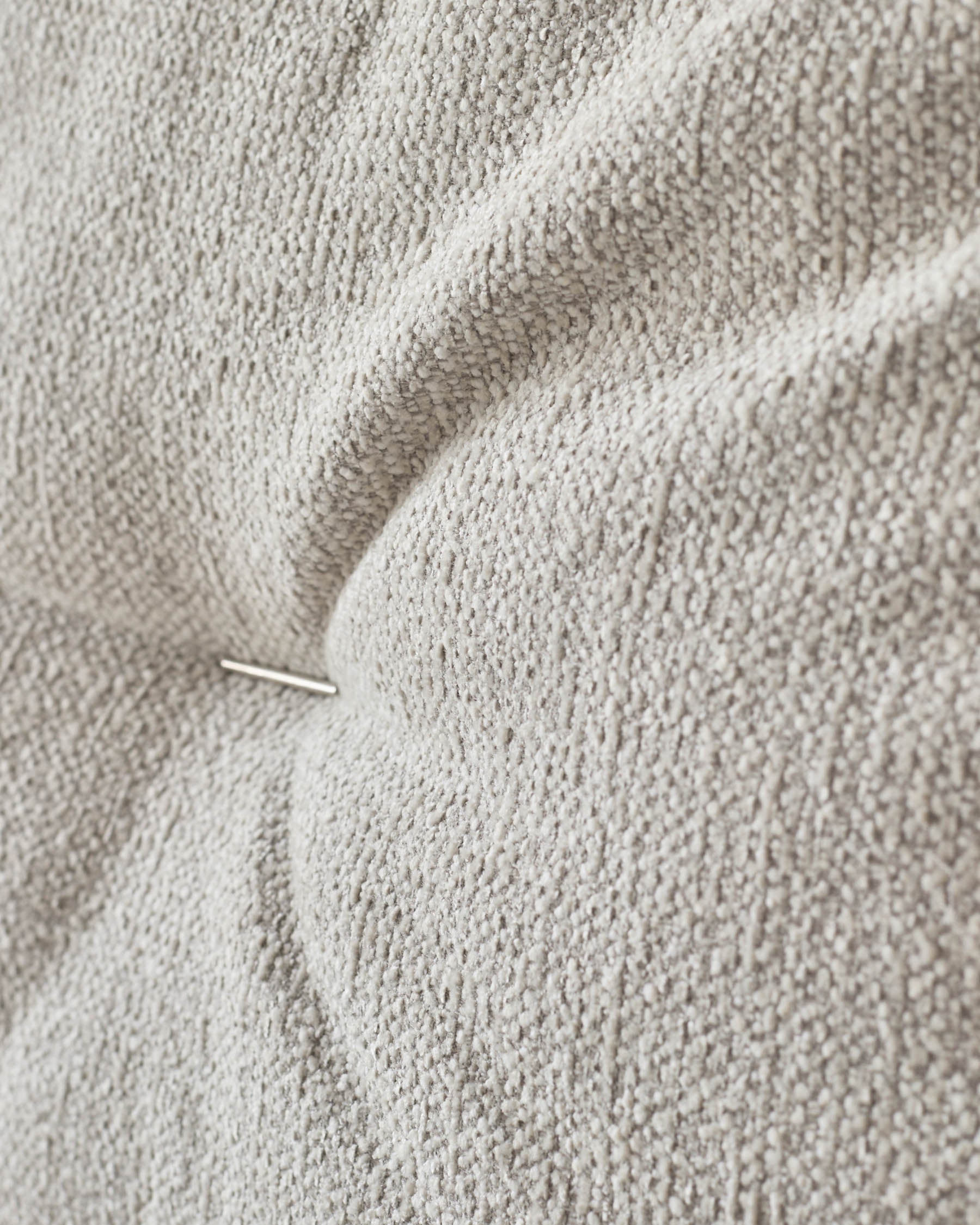 Close-up of textured, button-tufted upholstery in a neutral grey, featuring a visible pin for a detailed look.
