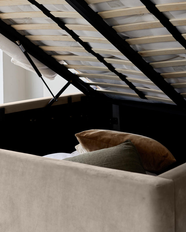 A beige ottoman bed with a sprung slatted base is lifted to reveal underbed storage with pillows.