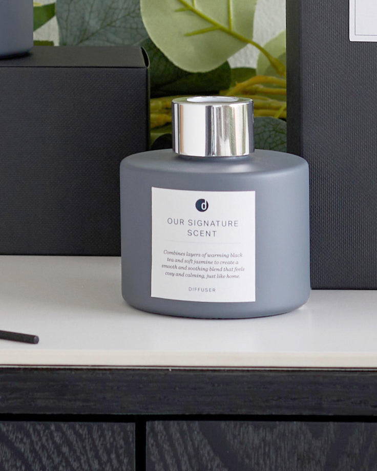 A light grey diffuser with a silver top sits on a dark wooden surface next to matching boxes.
