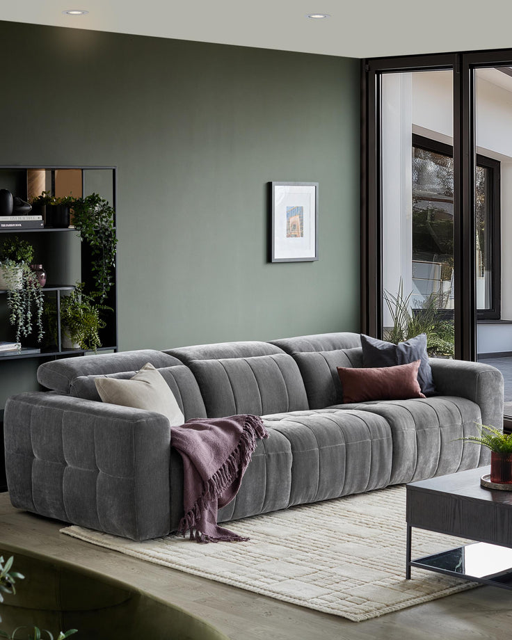 A large grey modular sofa with a coffee table, blanket, and pillows. There is a bookcase with plants in the background.
