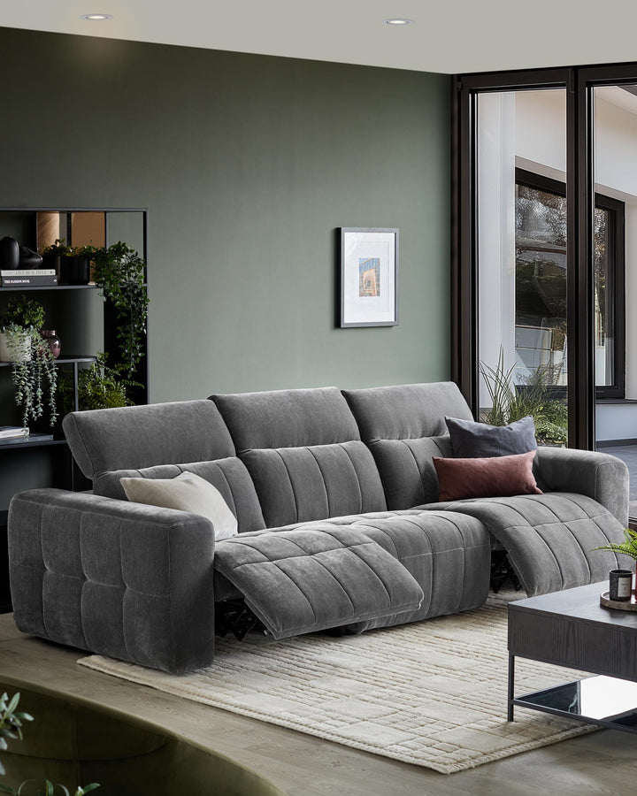 A grey, reclining sofa with cushions on a patterned rug, alongside a dark wood coffee table and bookshelf.