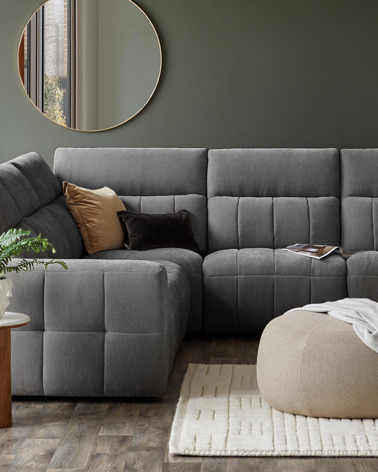 Stylish gray sectional sofa with plush cushions, a beige pouf, and a cozy white rug, complemented by a round mirror and greenery.