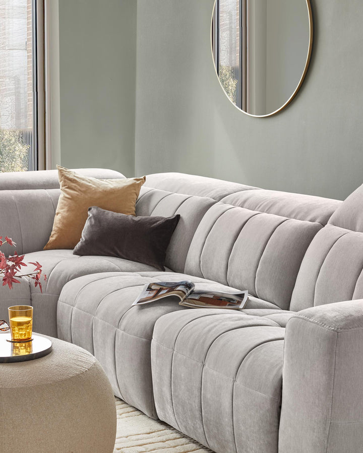 A plush, light gray sectional sofa with textured cushions, accented by decorative pillows and a round side table with a glass.