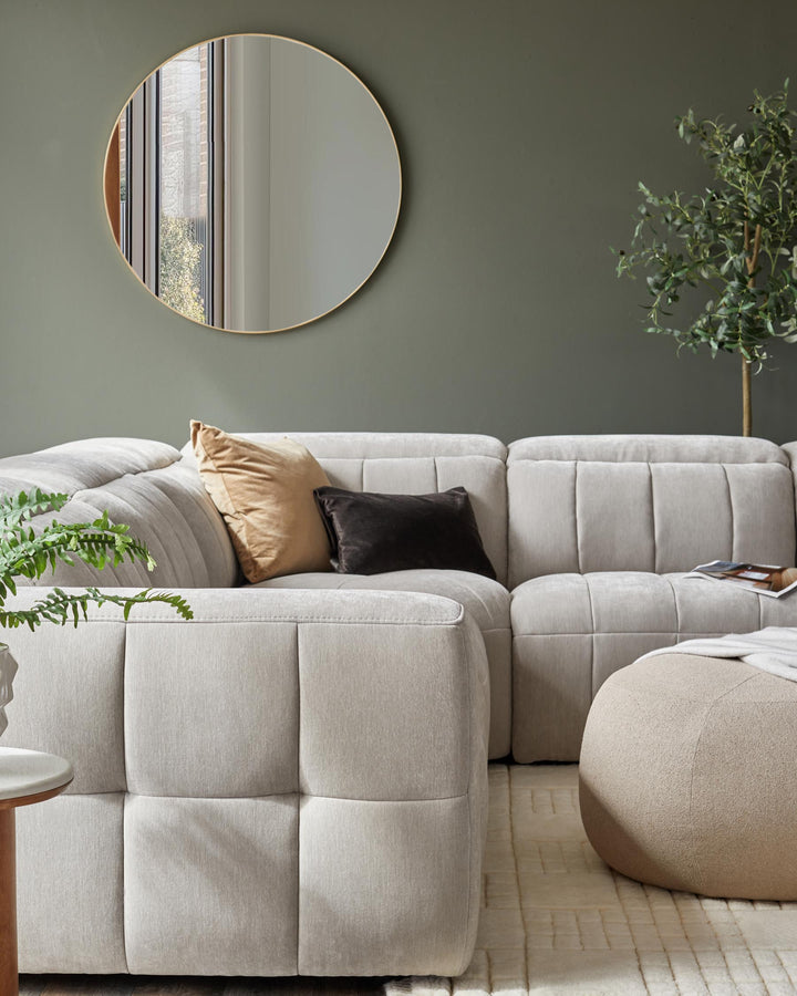 A cozy sectional sofa with soft cushions, a round wall mirror, a beige pouf, and a small side table with a plant.