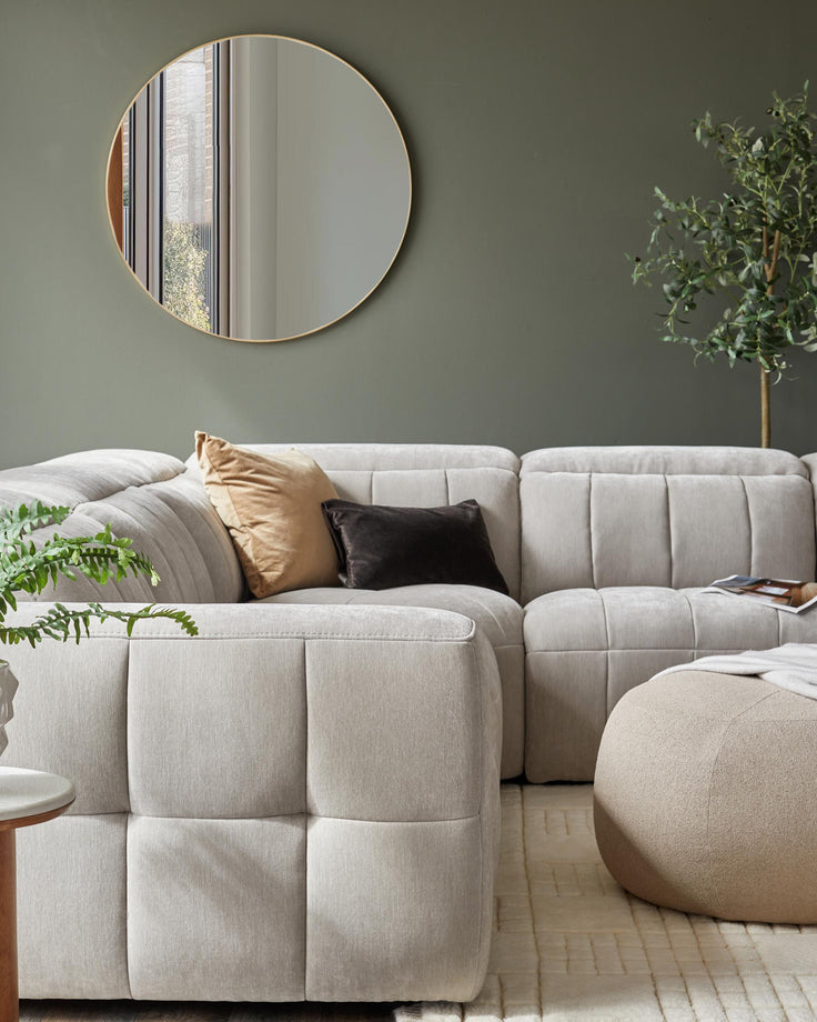A cozy sectional sofa with soft cushions, a round wall mirror, a beige pouf, and a small side table with a plant.