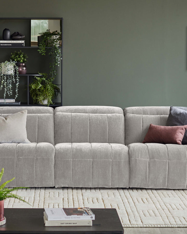 A light grey modular sofa with cushions, and a black shelving unit with books and pot plants, against a dark green wall.
