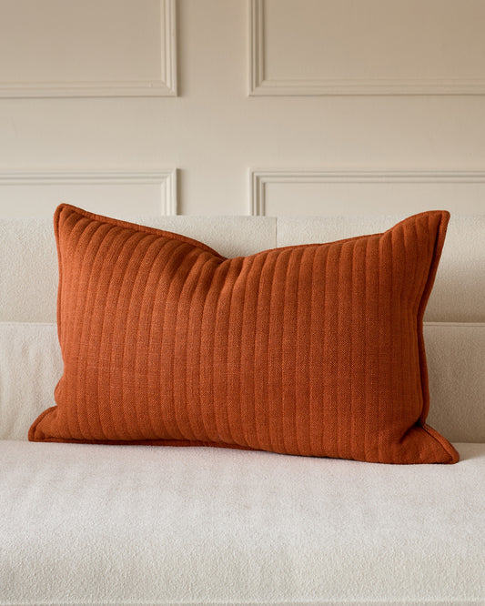 Rust-colored rectangular cushion with ribbed texture, displayed on a cream upholstered sofa against a paneled wall.