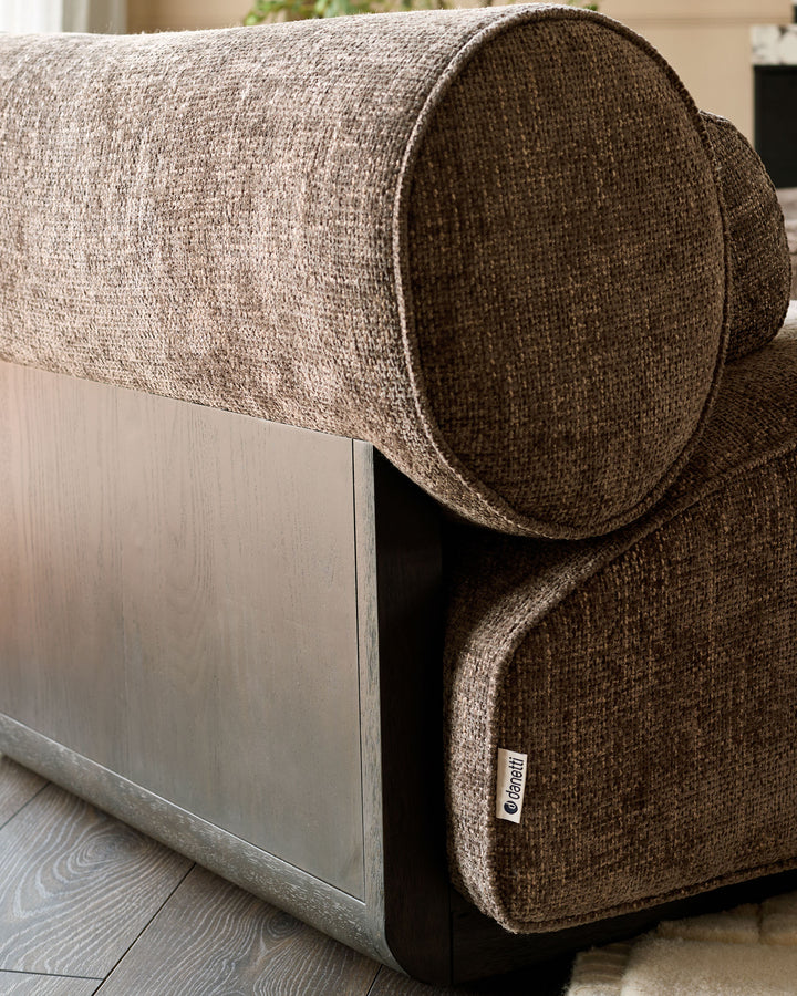 Sofia 2-seater sofa in mocha fabric, shown from side angle with cylindrical bolster pillow and wooden base detail.