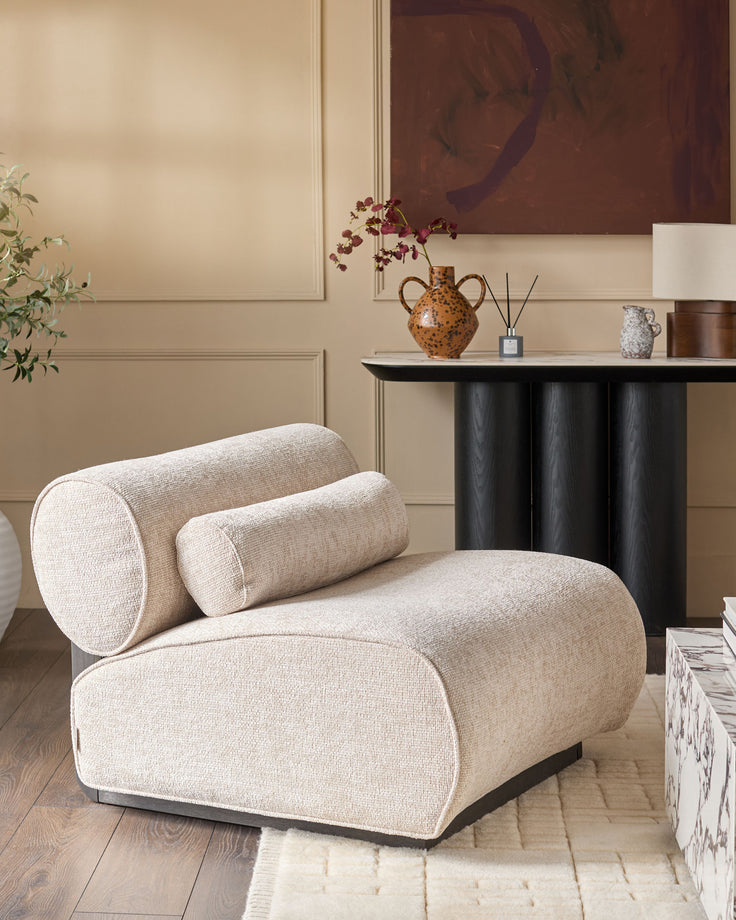 Sofia accent chair in natural fabric with cylindrical bolster pillows, curved design, and dark base in modern living room setting.