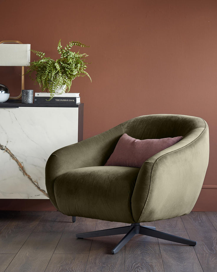 Olive green velvet swivel chair with a mauve cushion, sat on a dark wooden floor. A cabinet sits in the background.