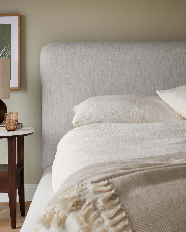 Bedroom with a fabric headboard, linen bedding and a dark wood bedside table topped with a lamp and a glass.