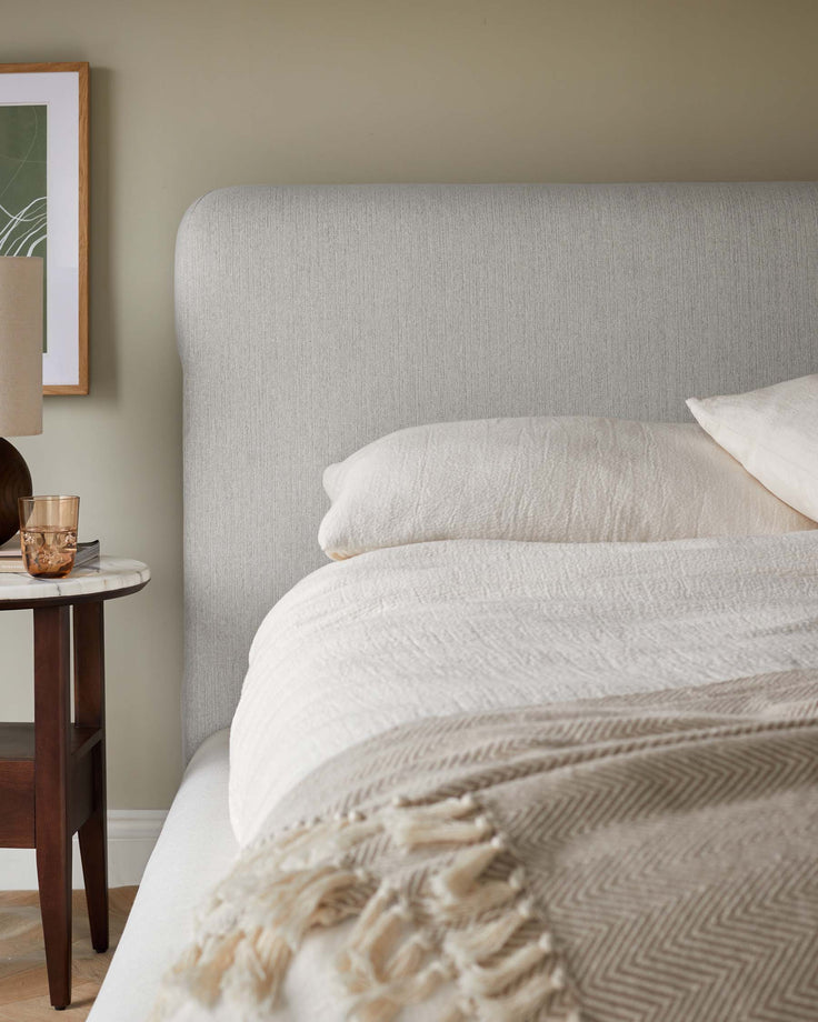 Bedroom with a fabric headboard, linen bedding and a dark wood bedside table topped with a lamp and a glass.