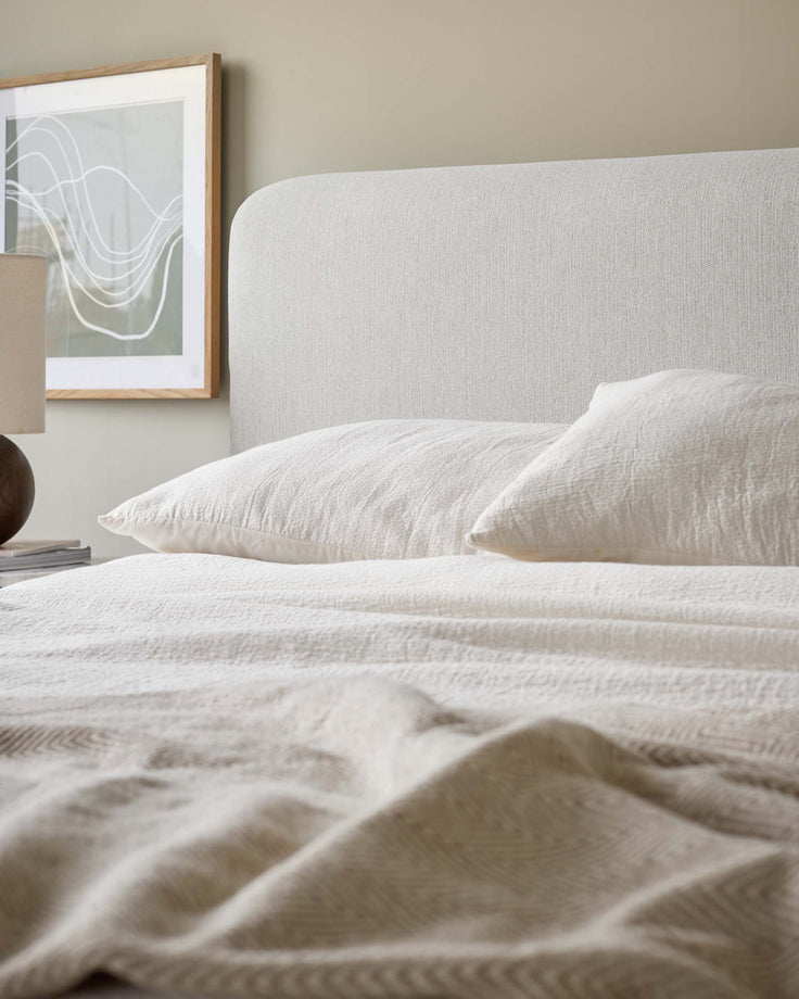 A made-up bed with a neutral colour scheme, featuring textured bedding, cushions, and a headboard.