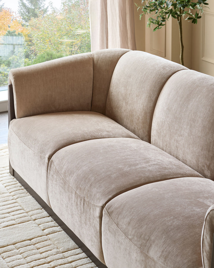 Teagan 3-seater sofa in champagne fabric, showing curved cushioned sections and dark base, positioned by a bright window.