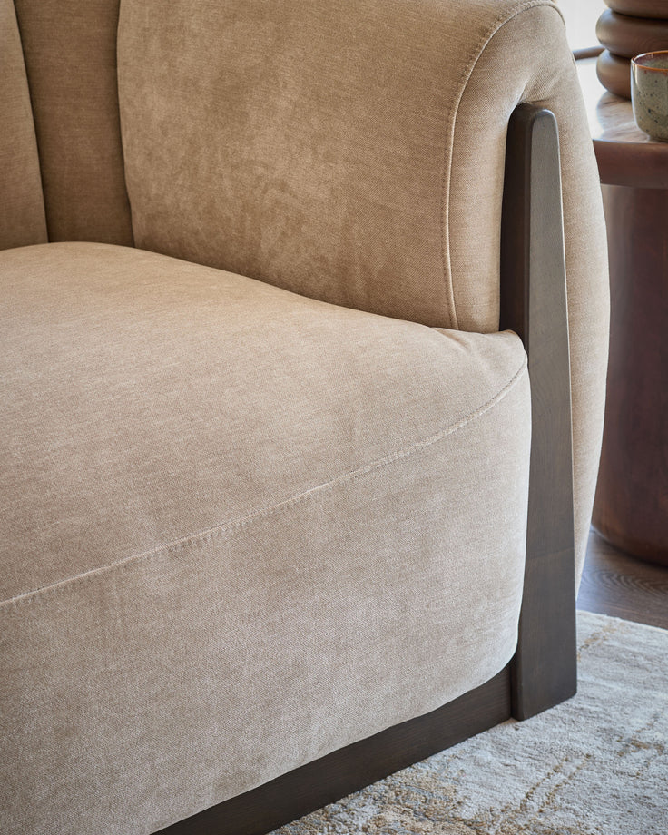 Teagan armchair in champagne fabric, side view showing curved seat, backrest, and Grey wooden arm detail.