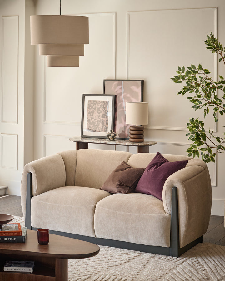 Teagan 2-seater sofa in champagne fabric with curved arms and dark base, styled with brown and burgundy pillows.