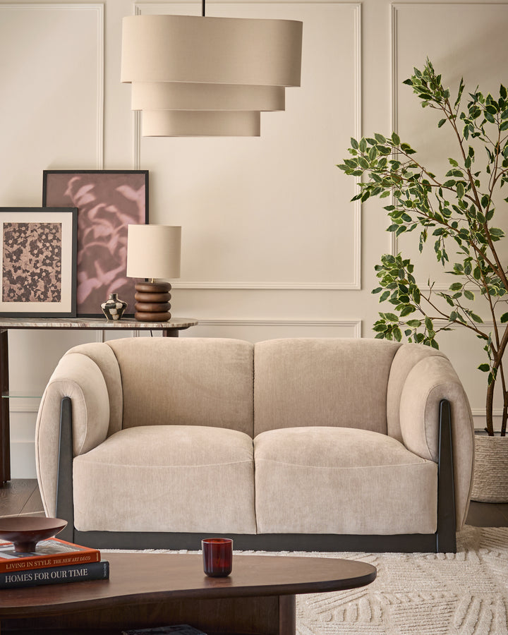 Teagan 2-seater sofa in champagne fabric with curved arms and dark wood base, displayed in a styled living room setting.