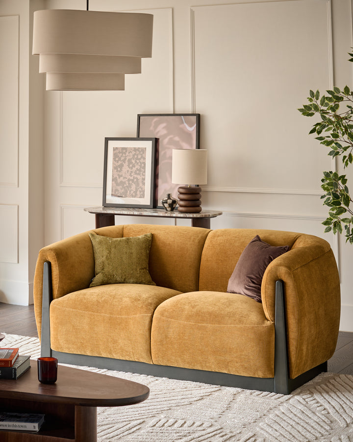 Teagan 2-seater sofa in ochre fabric with curved arms and dark frame, styled in a modern living room.