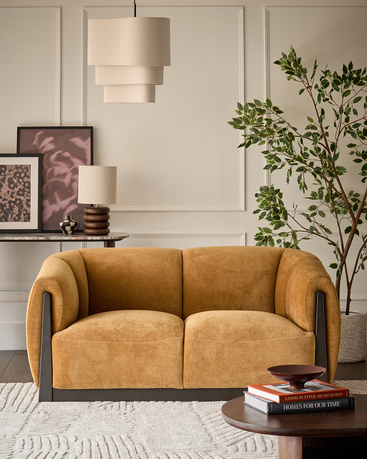 Teagan 2-seater sofa in ochre fabric with curved arms and dark wood base, positioned in a styled living room.