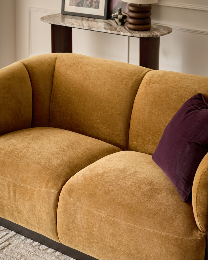 Teagan 2-seater sofa in ochre fabric with curved, rounded arms and back, styled with burgundy cushion.