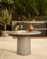 Grey pedestal dining table with round top set on a patio with wine glasses and terracotta vase.