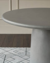 Grey round dining table with pedestal base, shown from side angle on geometric patterned floor.