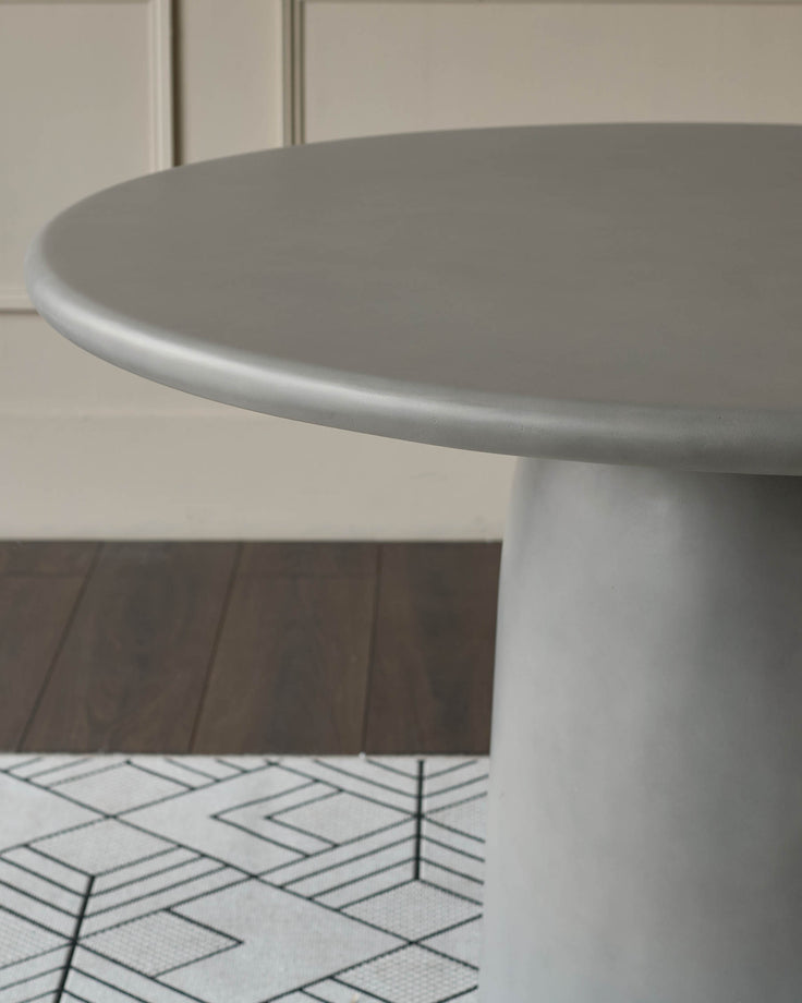 Grey round dining table with pedestal base, shown from side angle on geometric patterned floor.