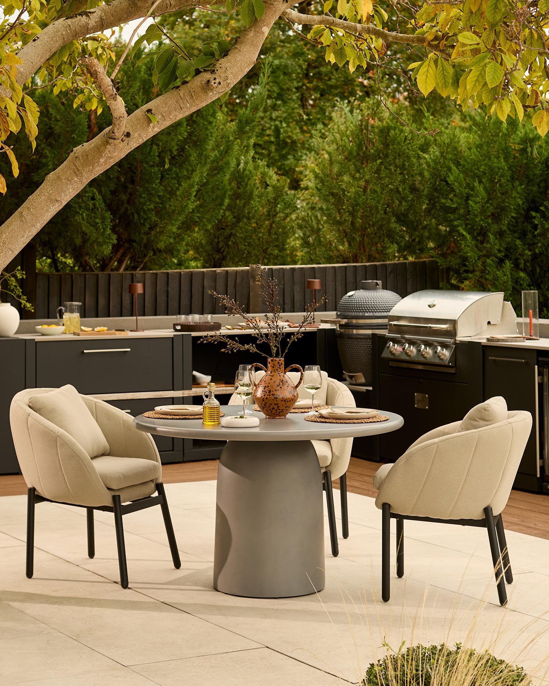 Dark grey cylindrical pedestal dining table set with two beige upholstered chairs in modern outdoor setting.