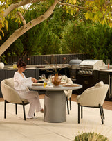 Tesso round grey dining table with four natural upholstered chairs in modern outdoor patio setting with kitchen area.