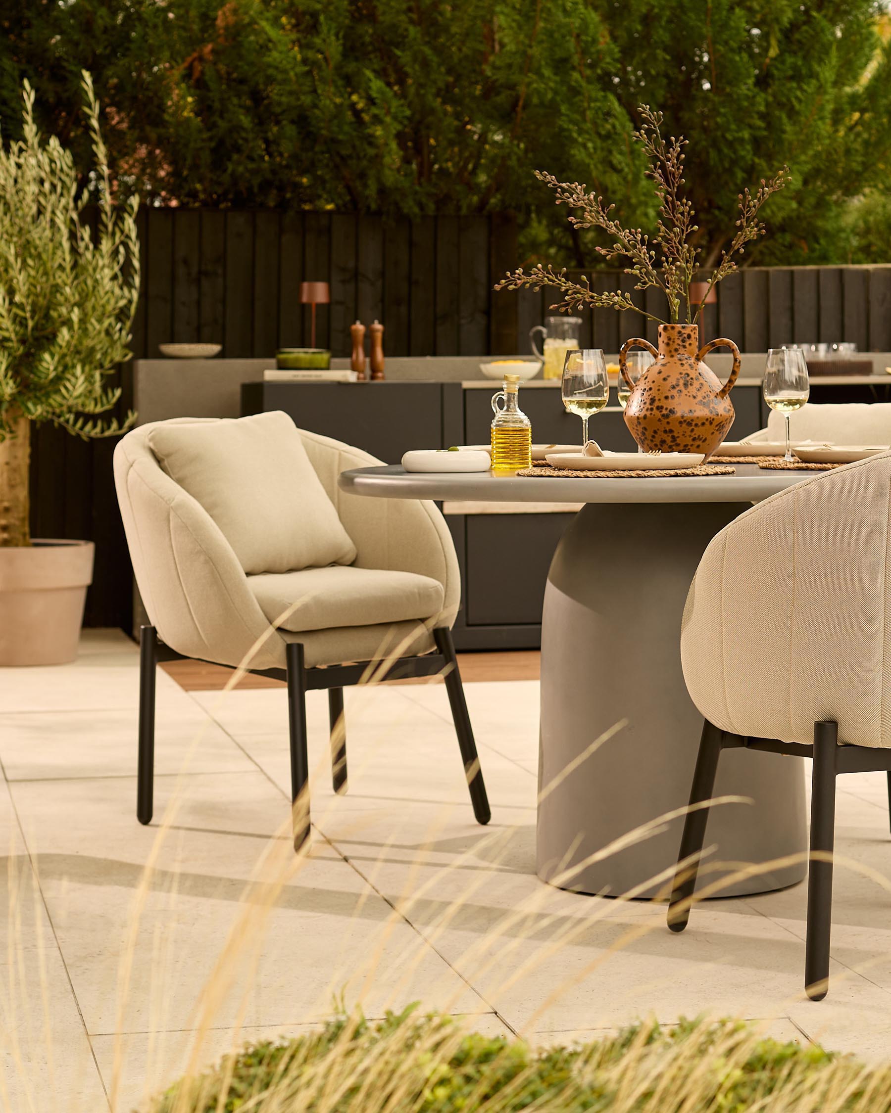 Tesso dark grey dining table with four natural beige Camilo chairs in modern garden setting with greenery and patio.