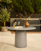 Grey cylindrical pedestal dining table with round top, styled in a modern outdoor patio setting with wine glasses and terracotta vase.