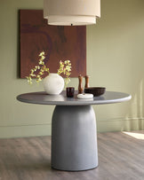 Dark grey round pedestal dining table styled with white vase, wooden accessories, and pendant lighting overhead.