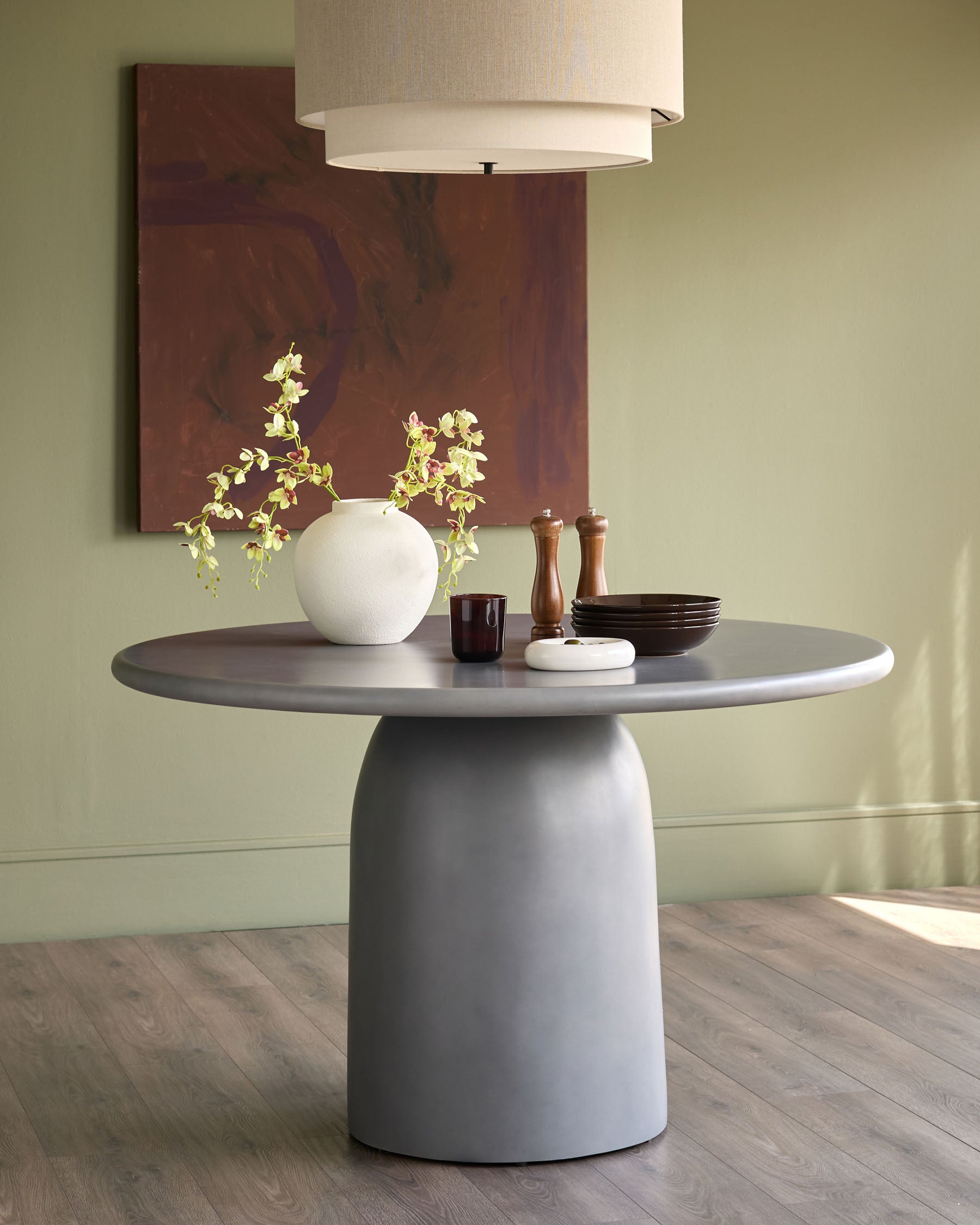 Dark grey round pedestal dining table styled with white vase, wooden accessories, and pendant lighting overhead.