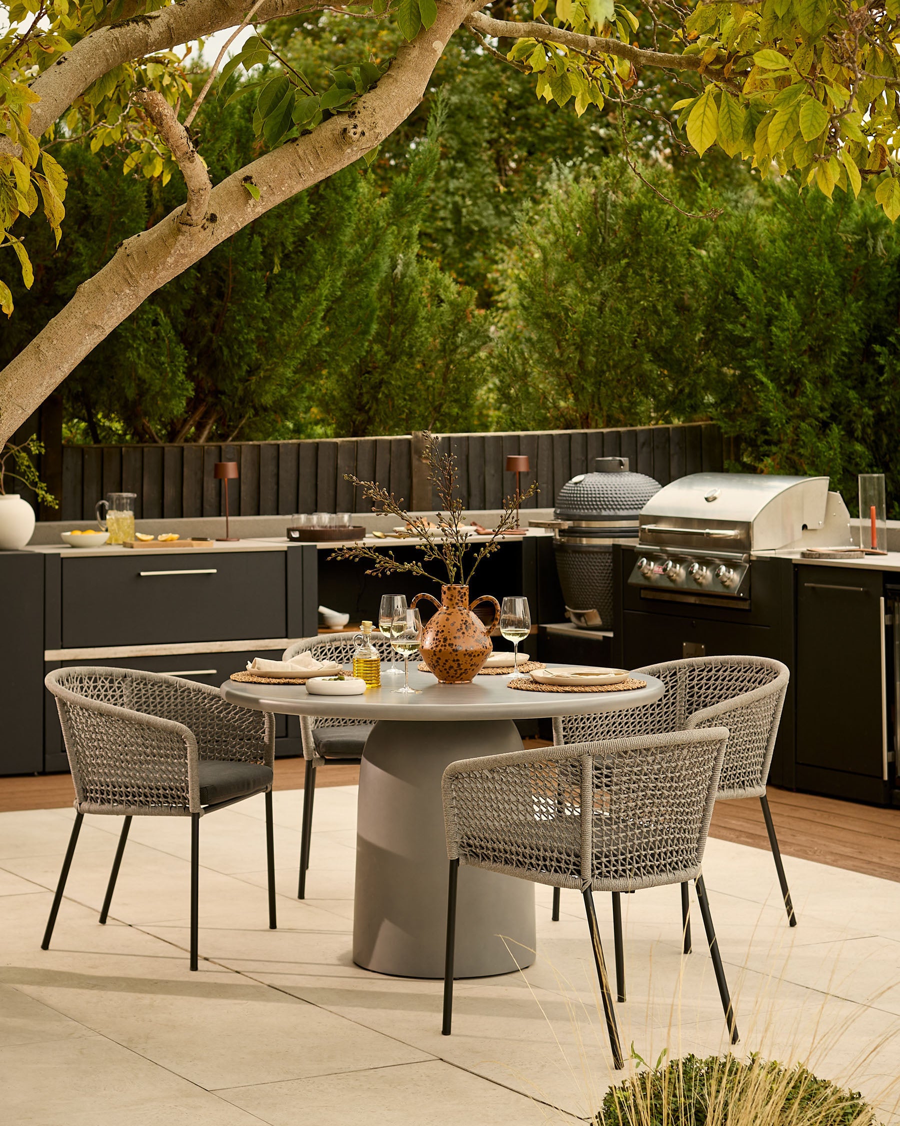 Tesso dark grey 4-seater round dining table styled in an outdoor kitchen setting with woven chairs and tree backdrop.
