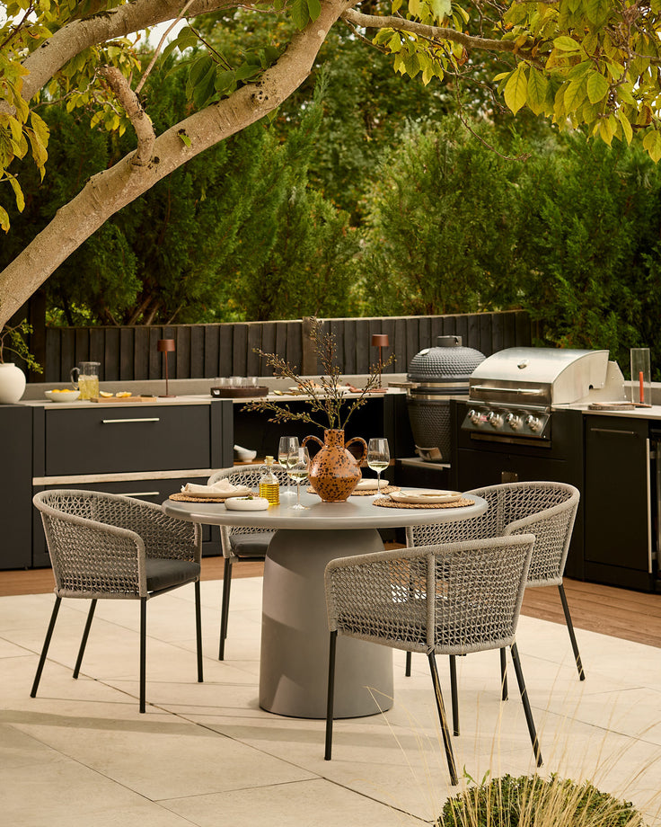 Tesso dark grey 4-seater round dining table styled in an outdoor kitchen setting with woven chairs and tree backdrop.