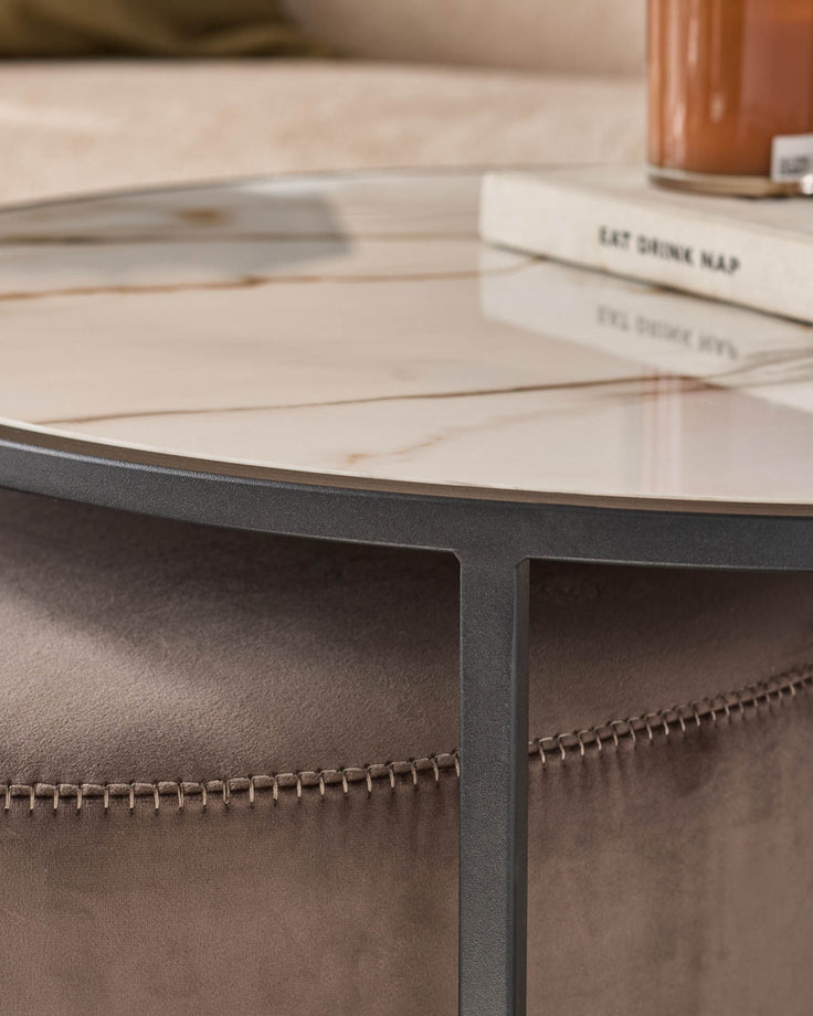 Round black coffee table with light wood top and mocha suede pouffe with stitched details, styled with books and decor.
