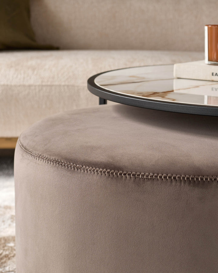 Mocha pouffe with decorative stitching detail, paired with black round coffee table in modern living room setting.