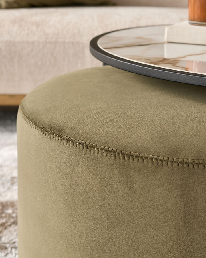 Olive green upholstered round pouffe with visible seam detail, paired with black-rimmed coffee table in neutral living room setting.